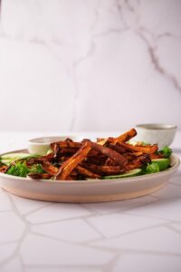 A pile of air fryer sweet potato fries on a plate, on top of a salad of greens, sliced avocado, and sliced cucumber. A bowl of dipping sauce sits on the side.