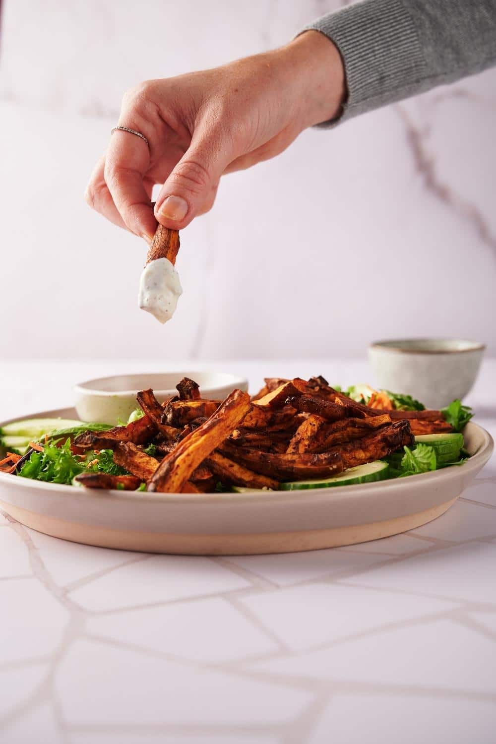 A hand dipping a sweet potato fry into a bowl of creamy dipping sauce. The sauce sits on a plate of sweet potato fries laid on a salad of greens and cucumber slices.