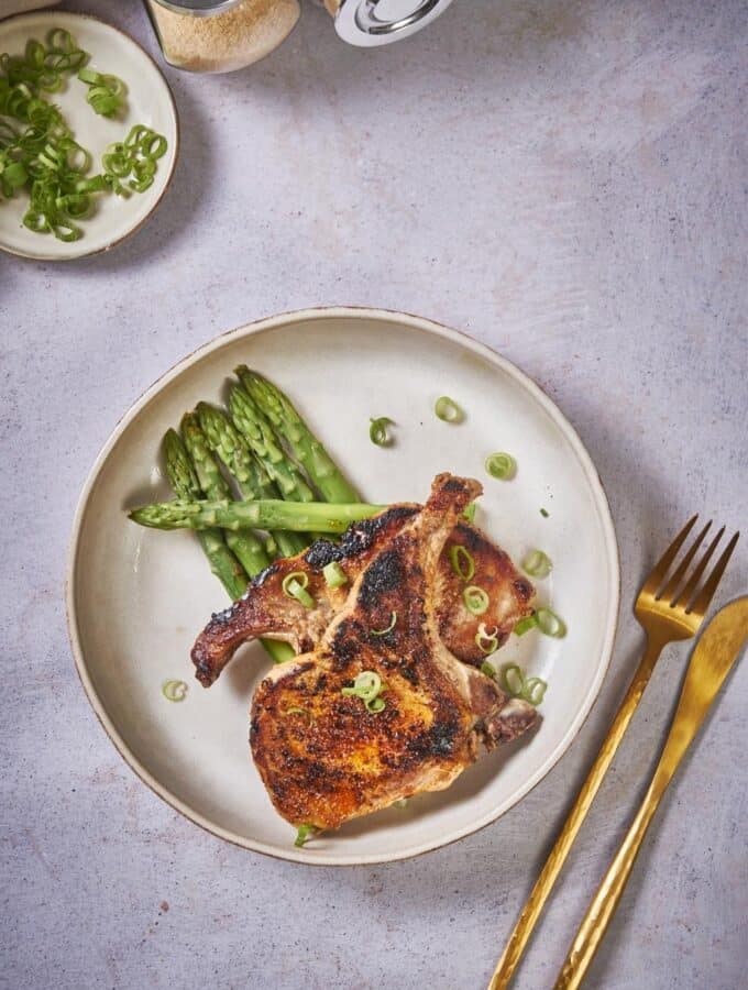 Two seared pork chops stacked on top of each other on a bed of asparagus. The pork chops are seasoned with chopped green onions. and are in an ecru colored plate. Next to the plate is a golden fork and a golden knife.