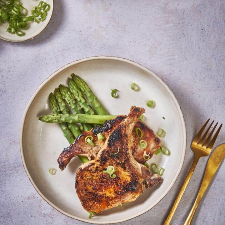 Two seared pork chops stacked on top of each other on a bed of asparagus. The pork chops are seasoned with chopped green onions. and are in an ecru colored plate. Next to the plate is a golden fork and a golden knife.