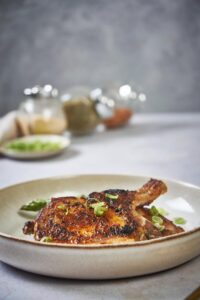 Two seared pork chops stacked on top of each other over cooked a bed of cooked aspargus in a deep ecru plate garnished with green onions.