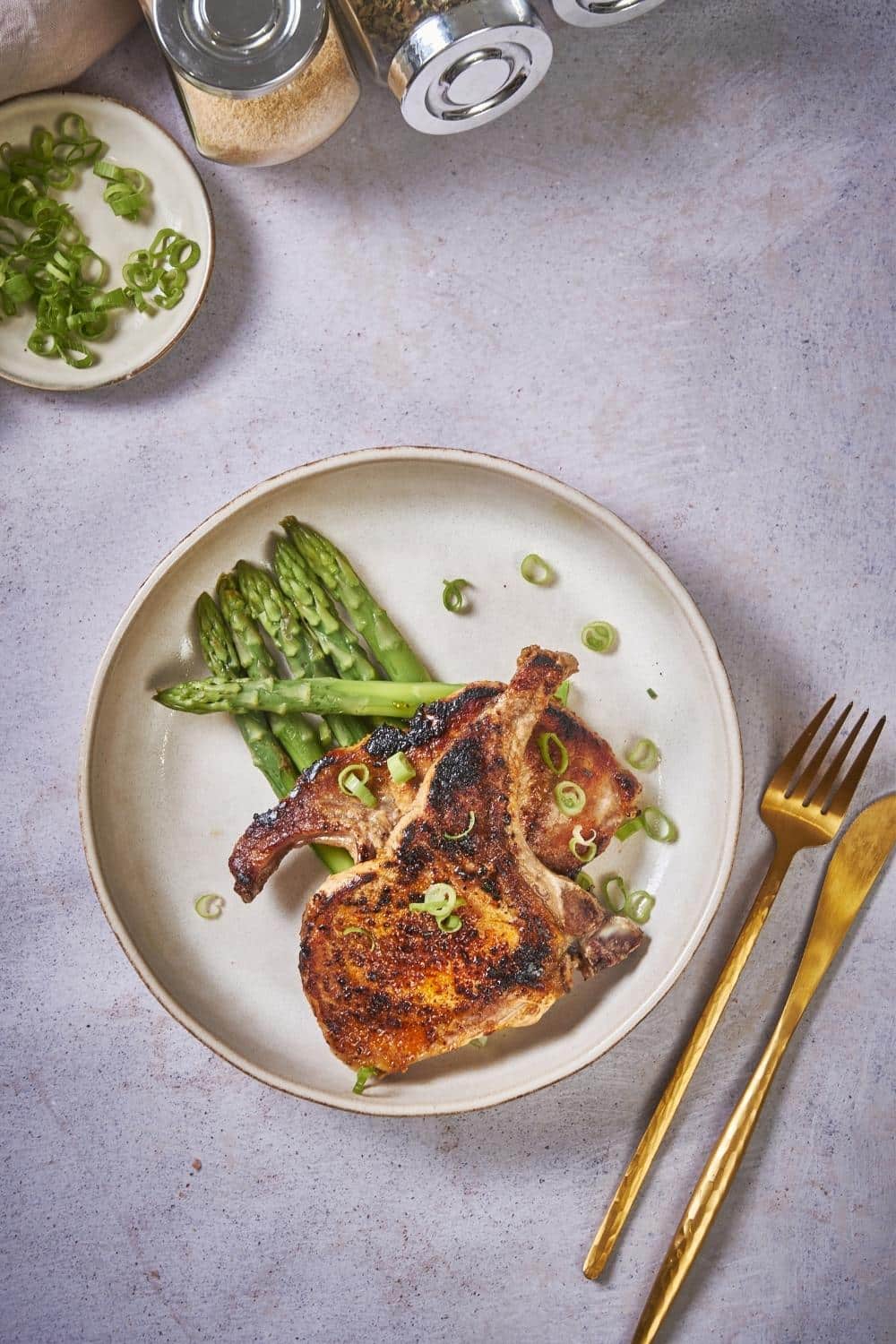 Two seared pork chops stacked on top of each other on a bed of asparagus. The pork chops are seasoned with chopped green onions. and are in an ecru colored plate. Next to the plate is a golden fork and a golden knife.