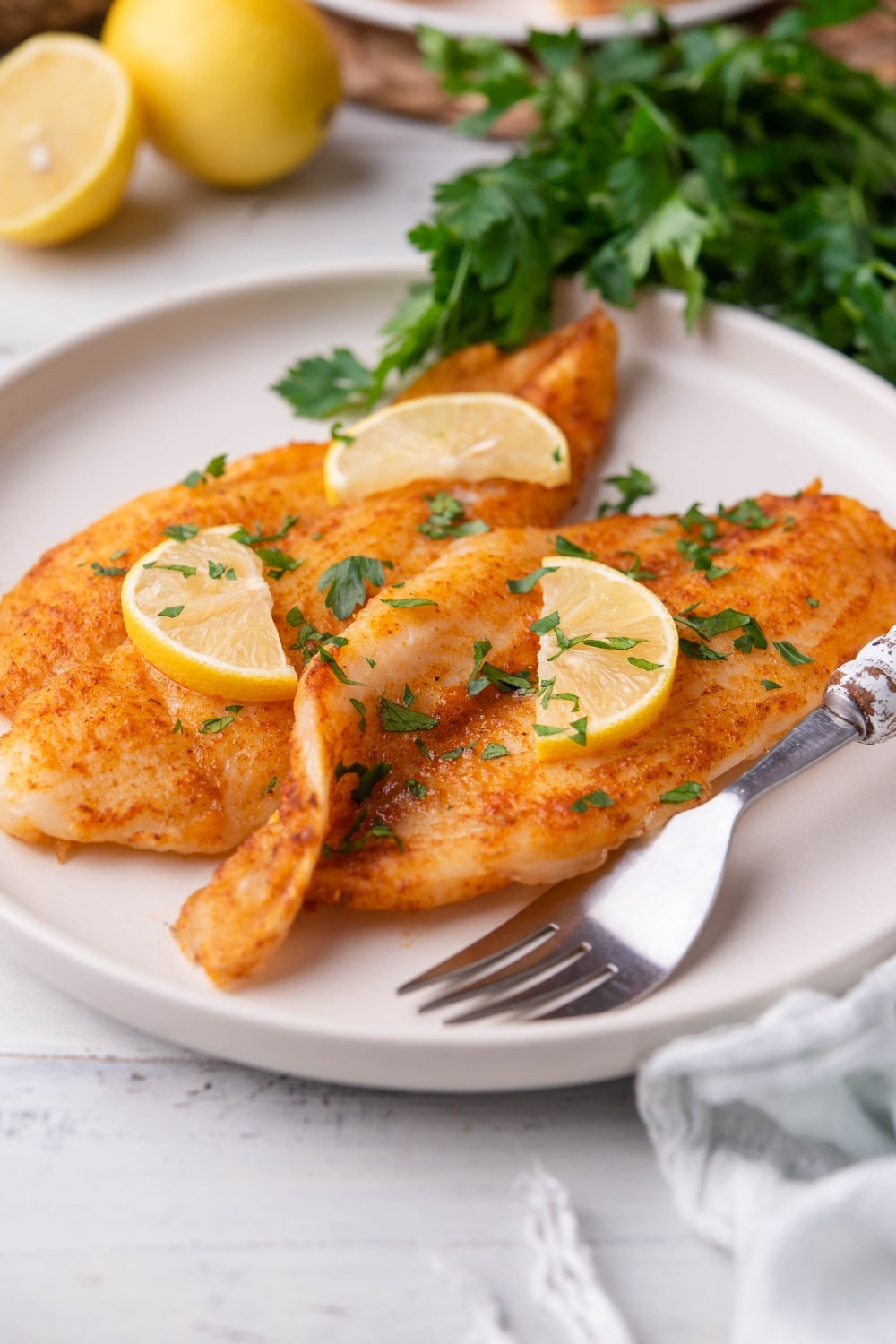 Air fried spiced tilapia fillets garnished with chopped parsley and lemon slices on a white plate with a fork. Surrounding the plate is a bundle of fresh parsley, lemons, and a tea towel.