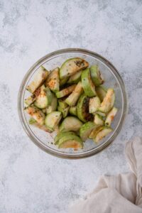 A glass bowl of sliced seasoned zucchini coated in olive oil, salt, pepper, garlic powder, onion powder, dried parsley, and paprika. Next to the bowl is a tea towel.