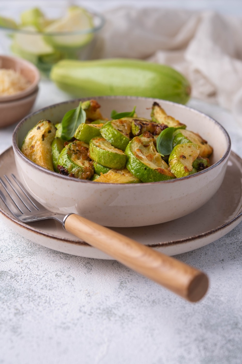 A bowl of air fried zucchini on a plate with a wooden handle fork. Behind the bowl is a large fresh zucchini, a glass bowl of sliced raw zucchini, and a small bowl of grated parmesan.