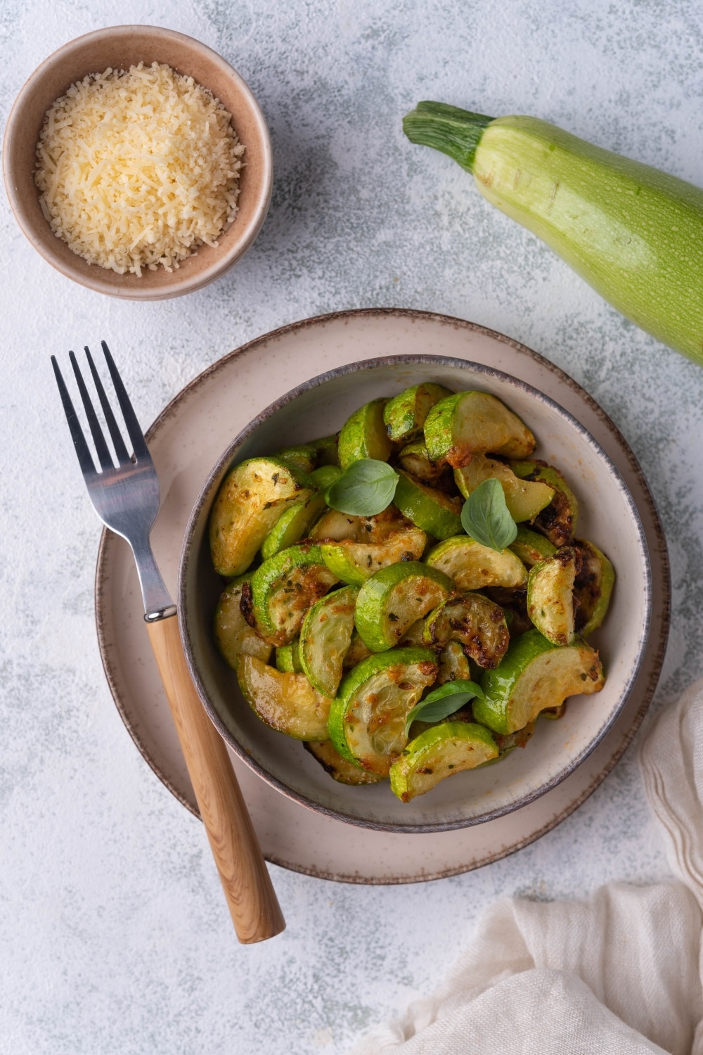 Top view of a bowl of air fried zucchini on a plate with a wooden handle fork. Surrounding the bowl is a fresh zucchini, a small bowl of grated parmesan, and a tea towel.