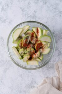 A glass bowl of sliced zucchini with unmixed herbs and seasonings on top. A tea towel is next to the bowl.