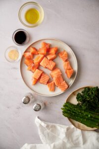 Raw cubed salmon on a plate. Surrounding the plate is a bowl of olive oil, smaller bowls of soy sauce and garlic powder, salt and pepper shakers, and a plate of asparagus and broccolini.