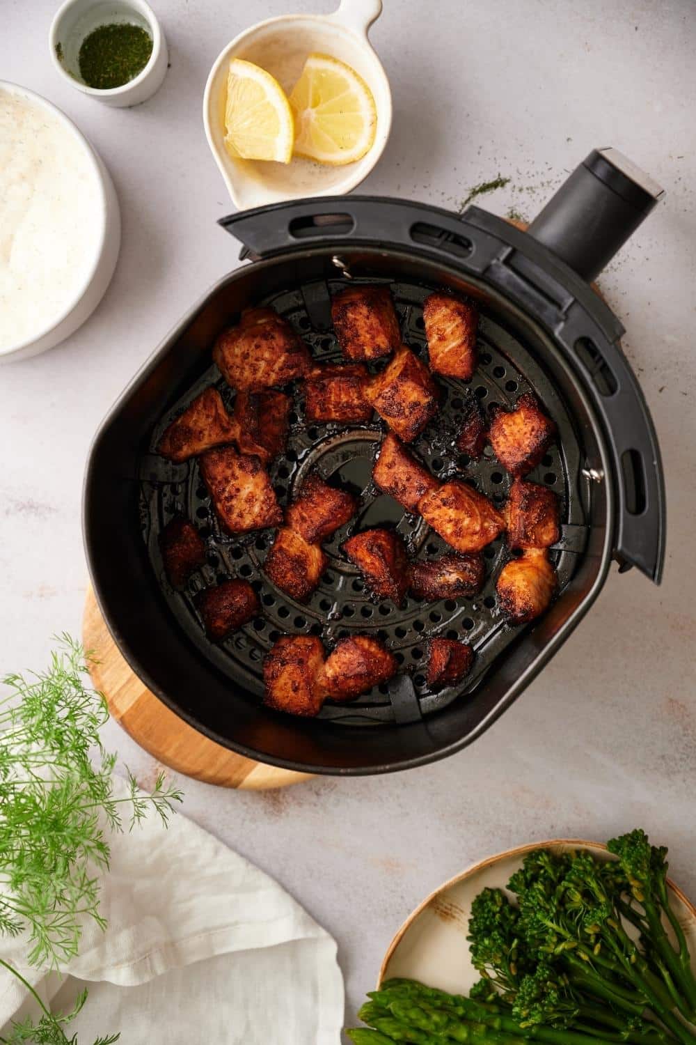 An air fryer basket filled with cooked salmon bites. The salmon bites are deep orange with browned edges. The basket is on top of a cutting board and it's surrounded by a small bowl of lemon wedges, a small bowl of chopped dill, a medium bowl of dill sauce, and part of a plate of asparagus and broccolini.