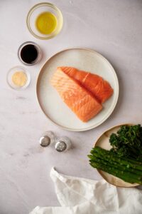 Two raw salmon fillets on a plate. Surrounding the plate is a bowl of olive oil, a small bowl of soy sauce, a small bowl of garlic powder, salt and pepper shakers, and a plate of steamed asparagus and broccolini.