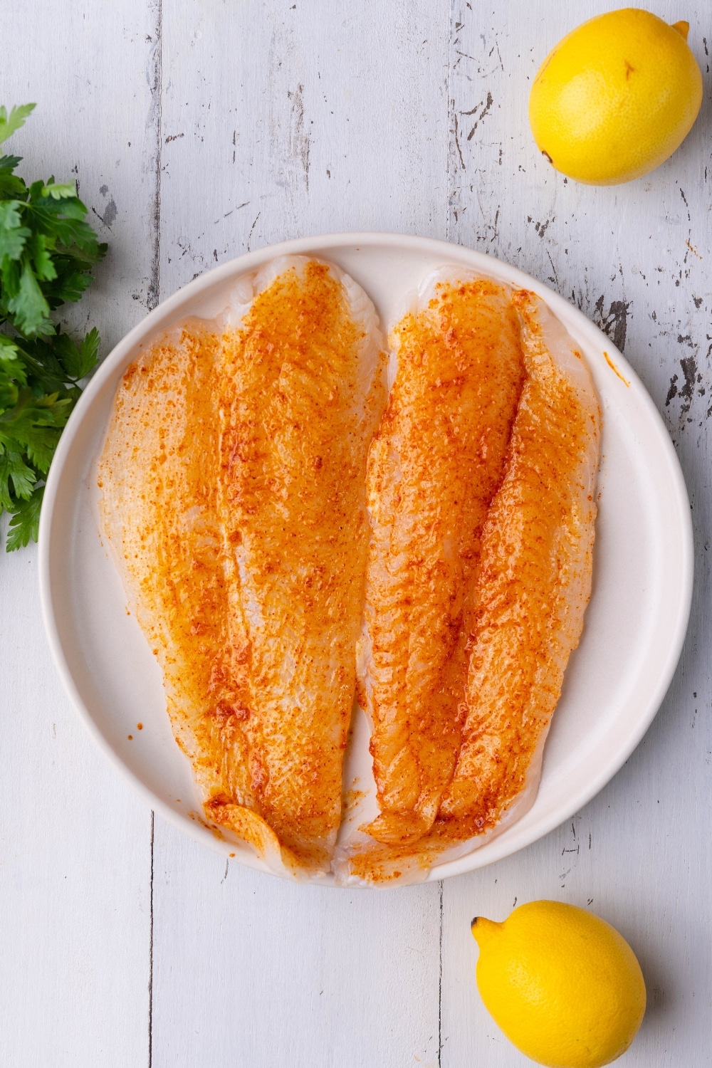 Seasoned raw tilapia fillets on a white plate. Surrounding the plate are lemons and a bundle of fresh parsley.