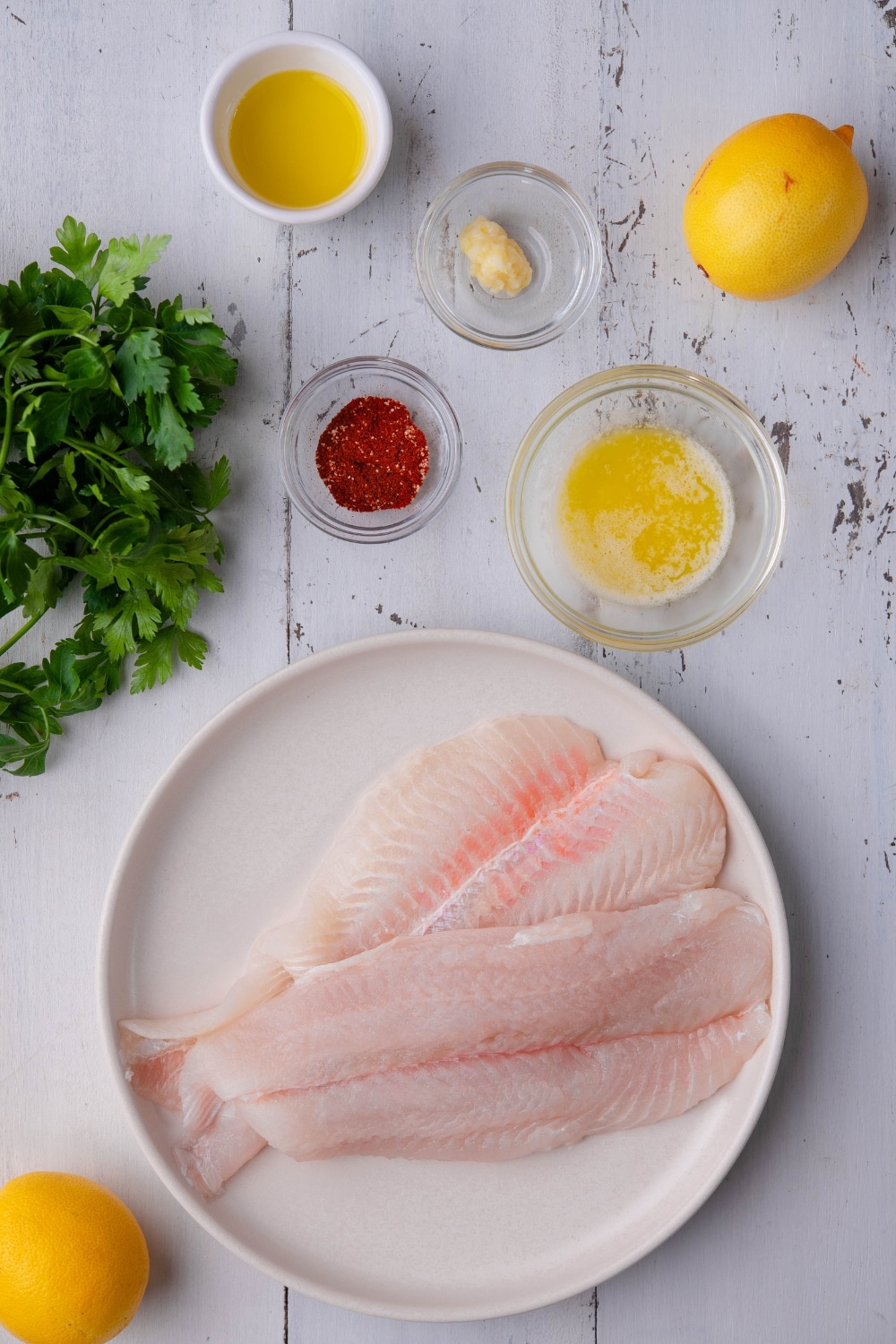 Two raw tilapia fillets on a white plate, whole lemons, a bunch of fresh parsley, and small bowls of olive oil, melted butter, garlic paste, and Cajun seasoning.