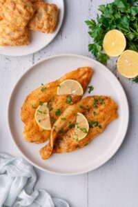 Two air fried tilapia fillets garnished with fresh chopped parsley and lemon slices. Surrounding the plate are more lemon slices on a bunch of fresh parsley, a plate of square hand pies, and a tea towel.