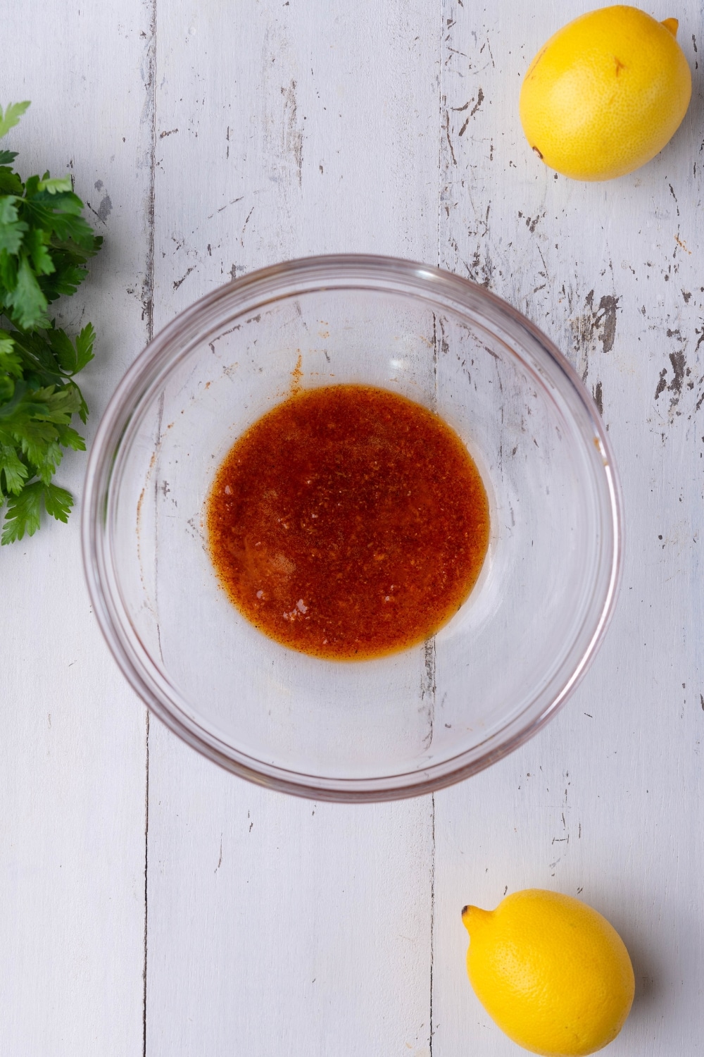 A glass bowl of butter and olive oil mixed with garlic paste and Cajun seasoning. Surrounding it are whole lemons and a bunch of fresh parsley.