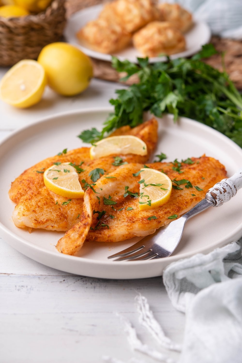 Two air fried tilapia fillets garnished with fresh chopped parsley and lemon slices on a white plate with a fork. Surrounding the plate is a bunch of fresh parsley, a plate of square hand pies, part of a small basket of lemons, and a tea towel.