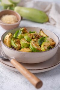 A bowl of air fried zucchini garnished with herbs on a plate with a wooden handle fork. Behind the bowl is a large fresh zucchini, a small bowl of grated parmesan, and a tea towel.