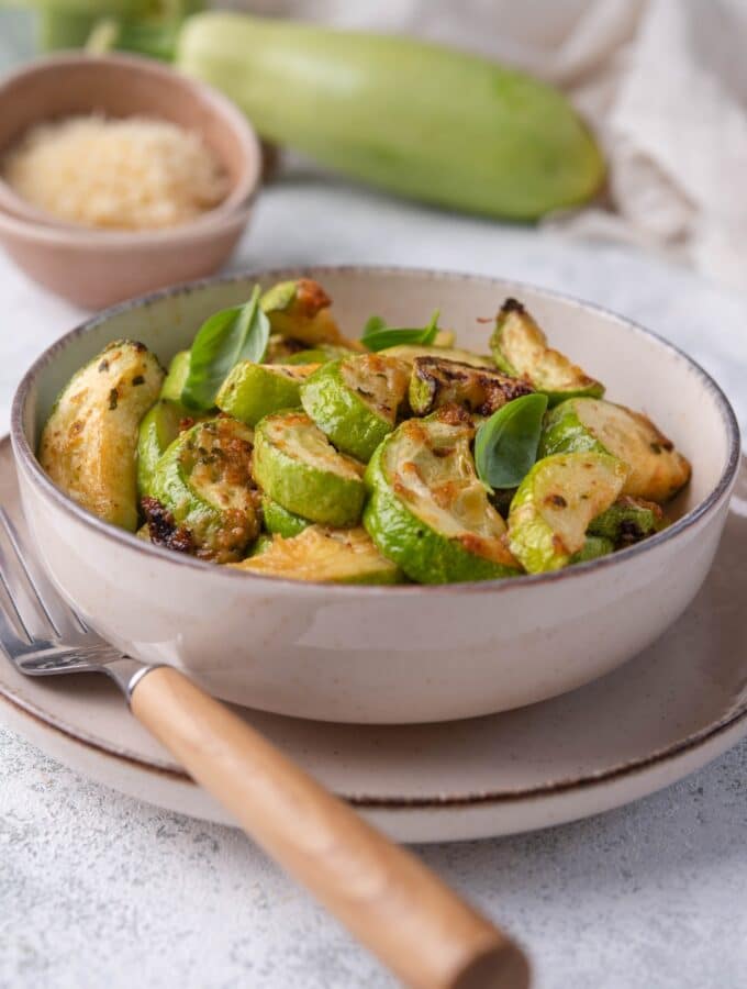 A bowl of air fried zucchini garnished with herbs on a plate with a wooden handle fork. Behind the bowl is a large fresh zucchini, a small bowl of grated parmesan, and a tea towel.