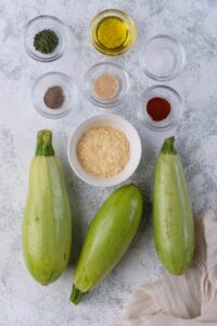 Three zucchinis, a medium bowl of grated parmesan, and small glass bowls of olive oil, garlic powder, dried parsley, paprika, salt, and pepper.