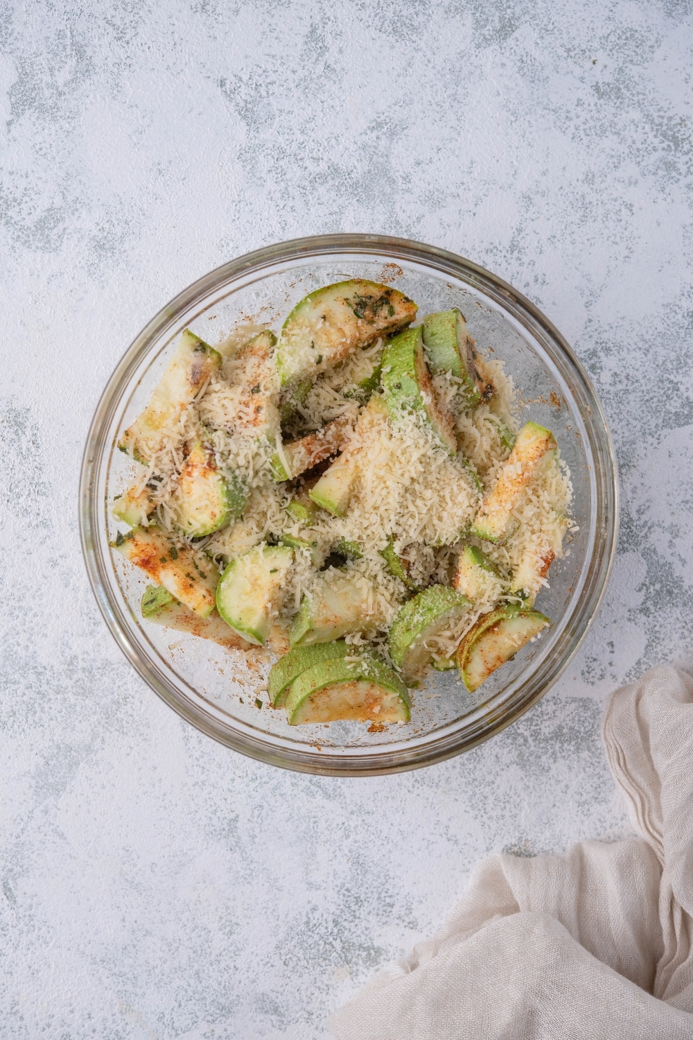 A glass bowl of sliced seasoned zucchini coated in herbs, spices, and grated parmesan. Next to the bowl is a tea towel.