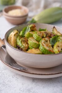 Close up of bowl of air fried zucchini on a plate with a wooden handle fork. The zucchini is garnished with fresh herbs. Behind the bowl is a large fresh zucchini and a small bowl of grated parmesan.