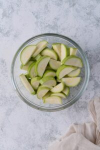 A glass bowl of sliced fresh zucchini next to a tea towel.