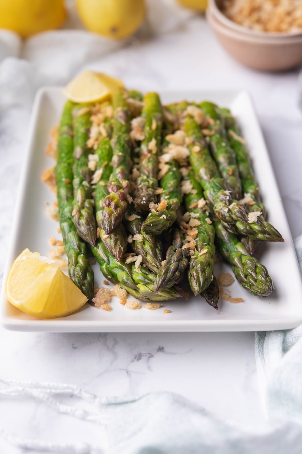 Sauteed asparagus garnished with panko breadcrumbs and served with lemon wedges on a rectangle white plate.