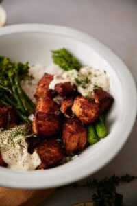 Close up of air fryer salmon cubes in a bowl. The salmon is in a bowl of white rice with asparagus and broccolini with two dollops of dill sauce garnished with fresh chopped dill.