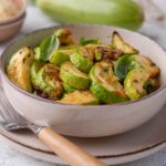 Closer look at air fried zucchini garnished with herbs in a bowl. The bowl is on plate with a wooden handle fork. Behind the bowl is a large fresh zucchini, a small bowl of grated parmesan, a glass bowl of zucchini slices, and a tea towel.