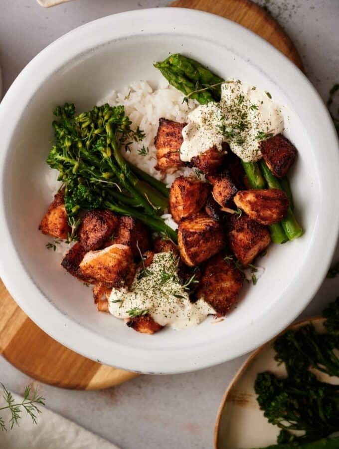 Air fryer salmon bites rice bowl. The bowl is filled with white rice topped with steamed asparagus and broccolini on either side with dollops of dill sauce and a generous serving of air fried salmon bites.