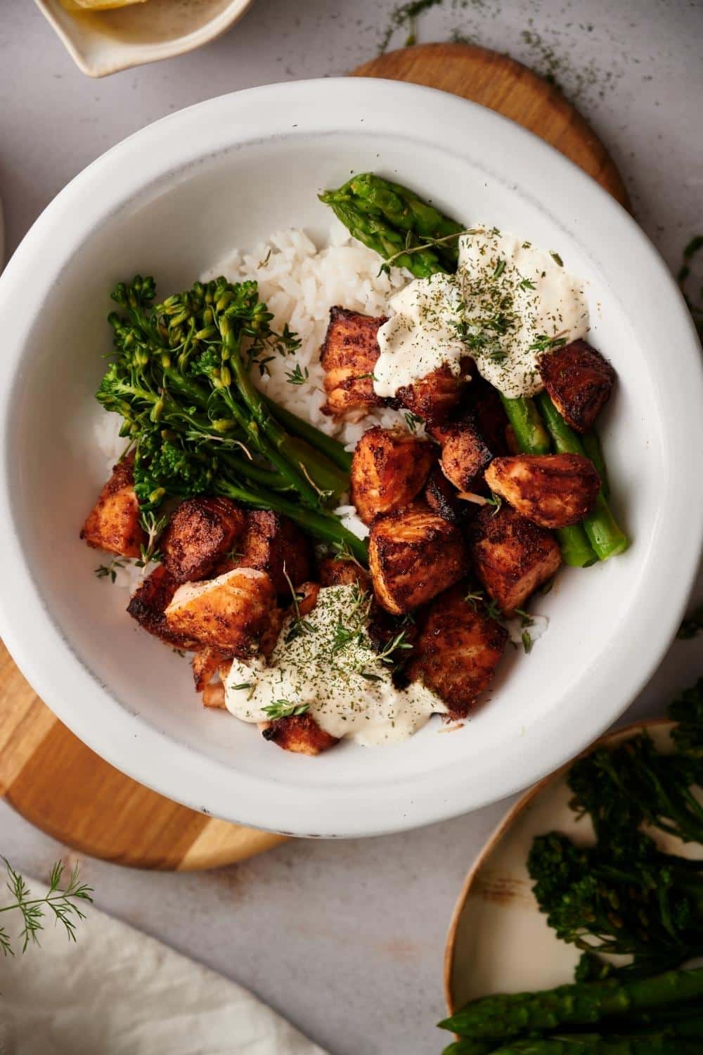 Air fryer salmon bites rice bowl. The bowl is filled with white rice topped with steamed asparagus and broccolini on either side with dollops of dill sauce and a generous serving of air fried salmon bites.