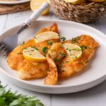 Air fried tilapia fillets garnished with fresh chopped parsley and lemon slices on a white plate with a fork. Behind it is a small basket of lemons, some that have been halved, and a plate of hand pies.