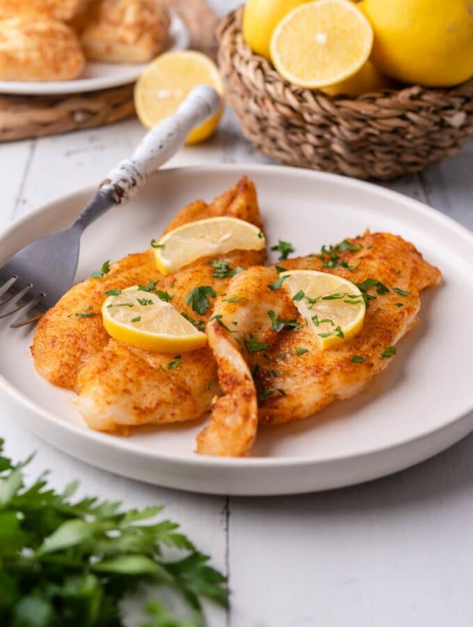 Air fried tilapia fillets garnished with fresh chopped parsley and lemon slices on a white plate with a fork. Behind it is a small basket of lemons, some that have been halved, and a plate of hand pies.