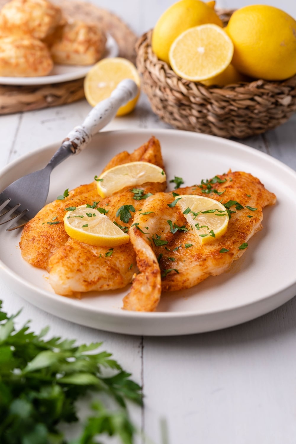 Air fried tilapia fillets garnished with fresh chopped parsley and lemon slices on a white plate with a fork. Behind it is a small basket of lemons, some that have been halved, and a plate of hand pies.