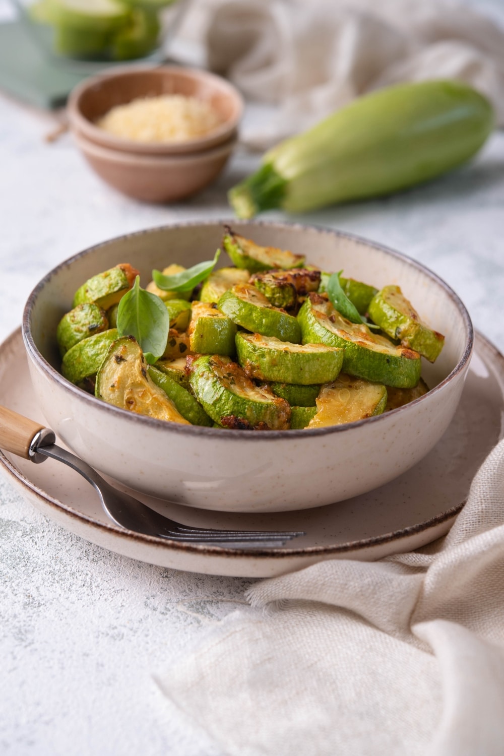 Air fried zucchini garnished with herbs in a bowl. The bowl is on plate with a wooden handle fork. Surrounding the plate is a fresh zucchini, a small bowl of grated parmesan, a glass bowl of zucchini slices, and a tea towel.