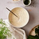 Fresh dill sauce in a medium sized white bowl with a metal spoon. Surrounding the bowl is a small bowl of lemon wedges, a ramekin filled with chopped dill, part of a plate of asparagus and broccolini, and a tea towel with fresh whole dill.