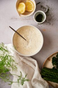 Fresh dill sauce in a medium sized white bowl with a metal spoon. Surrounding the bowl is a small bowl of lemon wedges, a ramekin filled with chopped dill, part of a plate of asparagus and broccolini, and a tea towel with fresh whole dill.