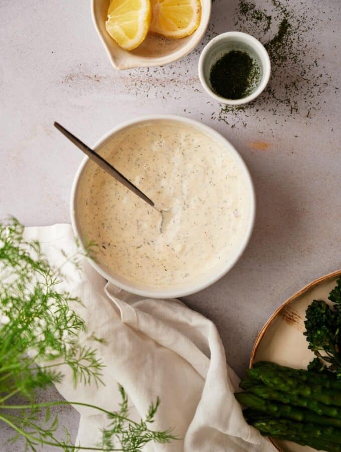 Fresh dill sauce in a medium sized white bowl with a metal spoon. Surrounding the bowl is a small bowl of lemon wedges, a ramekin filled with chopped dill, part of a plate of asparagus and broccolini, and a tea towel with fresh whole dill.