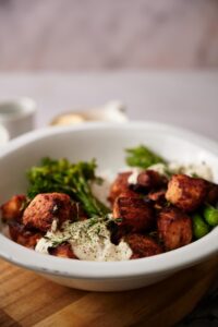 Air fried salmon bites in a white bowl. The salmon bites are served over white rice, steamed broccolini and asparagus, with two dollops of dill sauce garnished with fresh dill.