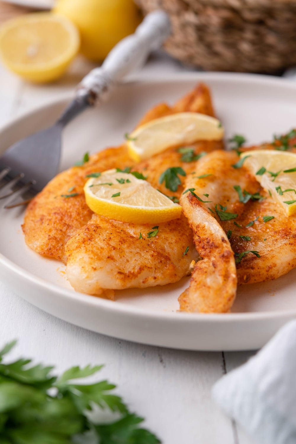 Close up of air fried tilapia fillets garnished with fresh chopped parsley and lemon slices on a white plate with a fork. Behind it is a small basket and some halved lemons.