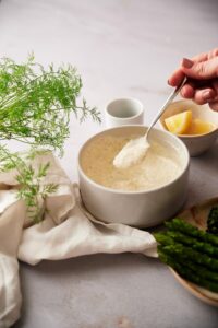 A spoon scooping fresh dill sauce from a medium sized white bowl. Behind the bowl is a small bowl of lemon wedges and a tiny bowl of chopped dill. Next to the bowl are a plate of asparagus and broccolini and fresh whole dill on top of a tea towel.