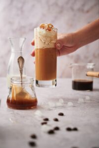 A hand picking up a glass of keto frappuccino. Surrounding it is a jar of caramel sauce, a carafe of almond milk, and a glass coffee pitcher filled with coffee.