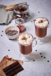 Two glasses of hot chocolate decorated with whipped cream and chocolate shavings. Surrounding the glasses is a jar of chopped chocolate, a small jar of maple syrup, a small container of chocolate chips, and a bar of baking chocolate resting on paper wrapper.