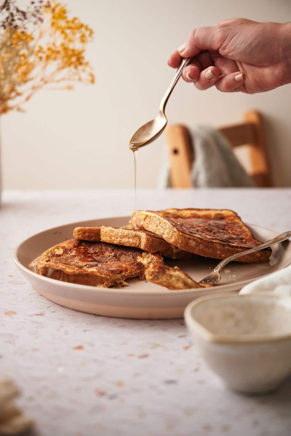 Three pieces of french toast arranged on a plate. A hand is pouring syrup over the toasts with a spoon and a fork with a piece of french toast is on the plate.