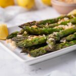 Close up of sauteed asparagus on a white rectangle plate. The asparagus is garnished with breadcrumbs and served with lemon wedges. In the back are whole lemons and a bowl of breadcrumbs.