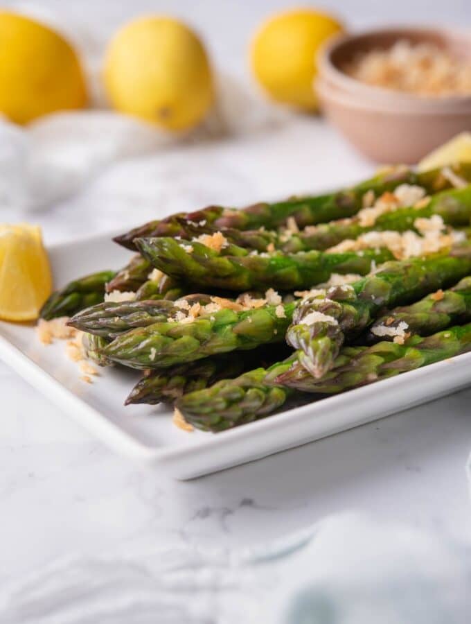 Close up of sauteed asparagus on a white rectangle plate. The asparagus is garnished with breadcrumbs and served with lemon wedges. In the back are whole lemons and a bowl of breadcrumbs.