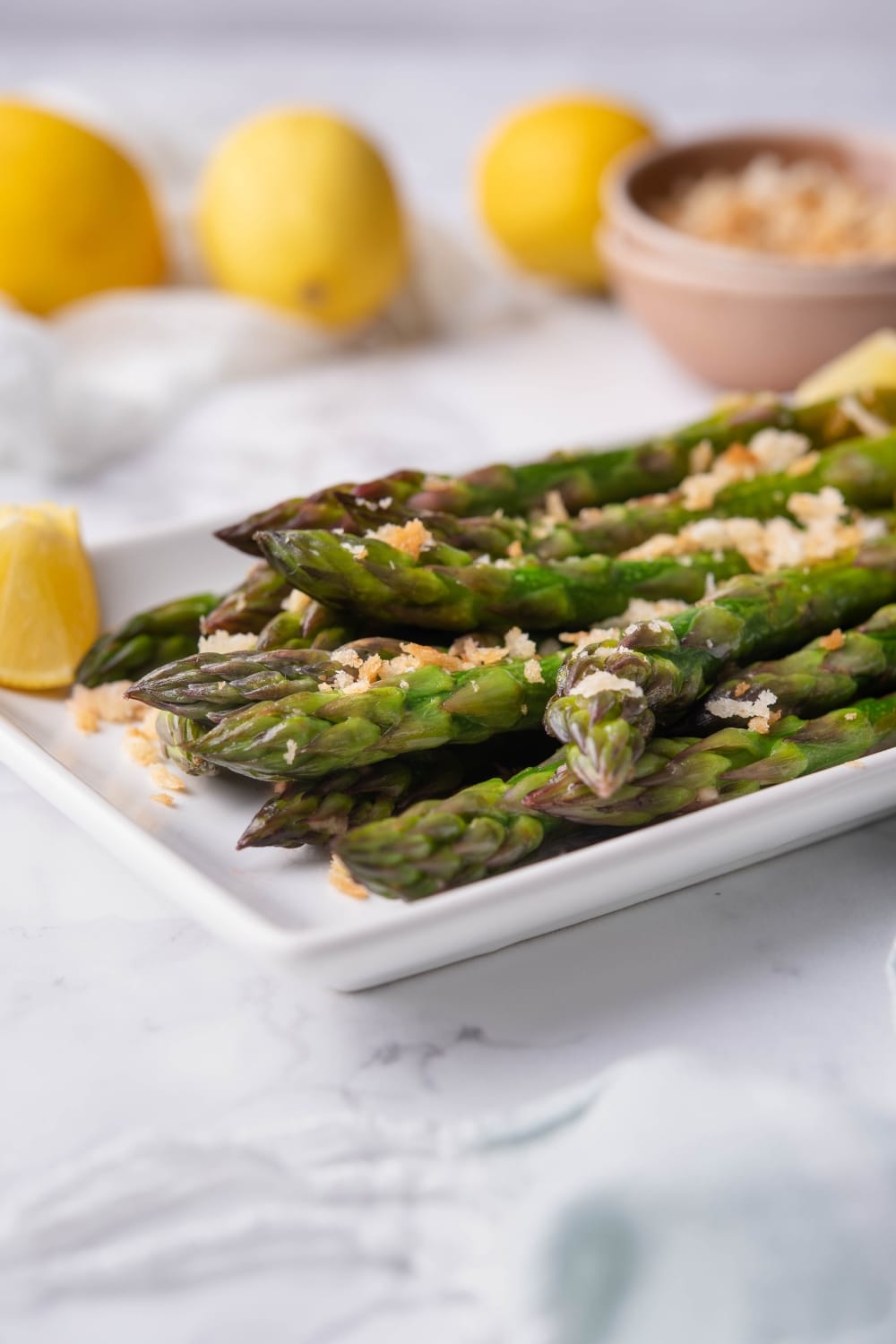 Close up of sauteed asparagus on a white rectangle plate. The asparagus is garnished with breadcrumbs and served with lemon wedges. In the back are whole lemons and a bowl of breadcrumbs.