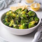 Sauteed broccoli with chili flakes and parmesan in a white bowl on top of a white plate. The bowl is garnished with a slice of lemon and more lemon slices can be seen in the background. A fork with a white handle is on the plate, next to the bowl.