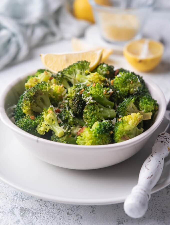 Sauteed broccoli with chili flakes and parmesan in a white bowl on top of a white plate. The bowl is garnished with a slice of lemon and more lemon slices can be seen in the background. A fork with a white handle is on the plate, next to the bowl.