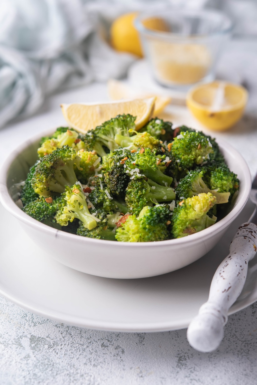 Sauteed broccoli with chili flakes and parmesan in a white bowl on top of a white plate. The bowl is garnished with a slice of lemon and more lemon slices can be seen in the background. A fork with a white handle is on the plate, next to the bowl.