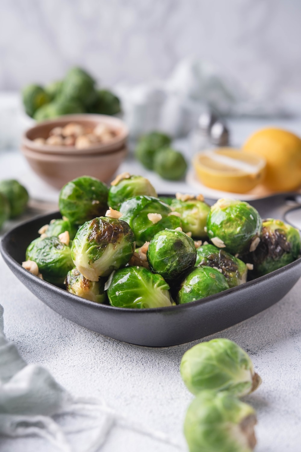 Sauteed brussels sprouts garnished with chopped hazlnuts in a small square cast iron skillet. Surrounding the pan are whole uncooked brussel sprouts, a bowl of hazelnuts, and a plate of lemon with half a lemon wedge.